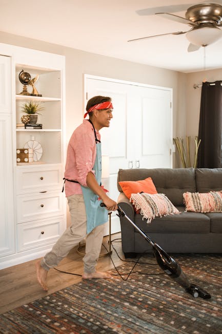 A person wearing orange work overalls is using a yellow and black vacuum cleaner with a long hose and a flat cleaning attachment to perform surface cleaning on a tiled floor in a modern living room. T