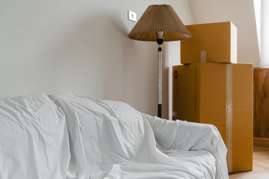A white fabric sofa covered with a protective sheet or slipcover, situated in a room with neutral-colored walls. To the right of the sofa, there are three stacked cardboard boxes of varying sizes, lik